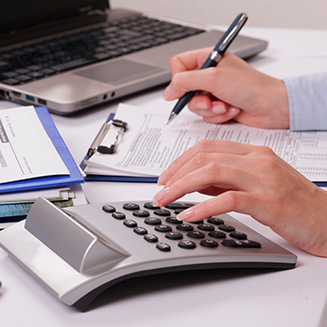 duas mãos utilizando calculadora e papel e caneta sobre uma mesa com um notebook ao fundo