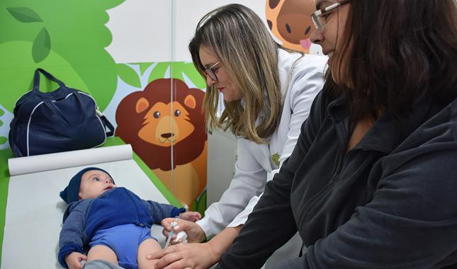 Em uma sala, um bebê está deitado em uma maca. Sua mãe e uma médica o acompanham na Clínica de Vacinas da Unimed Catanduva