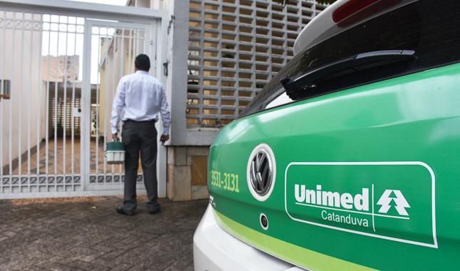 Em primeiro plano, a traseira de um carro com a marca da Unimed Catanduva. Ao fundo, colaborador está à porta de um beneficiário.