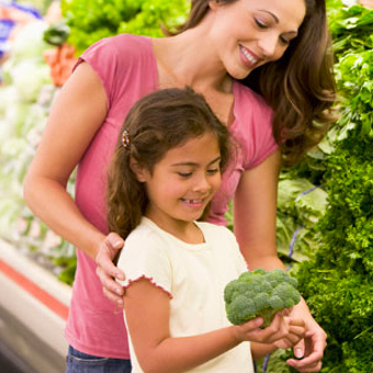 Imagem colorida. Mãe e filha pequena estão no setor de hortaliças do supermercado. A menina está sorrindo e segurando um brócolis.