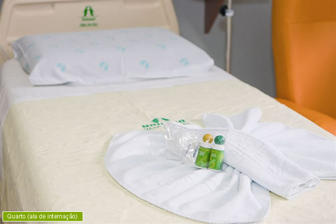 Imagem: Cama arrumada em um dos quartos de internação do Hospital Unimed Vale do Caí. Em cima dessa cama, está um travesseiro branco com o logotipo da Unimed e uma toalha branca com um kit de higienização.