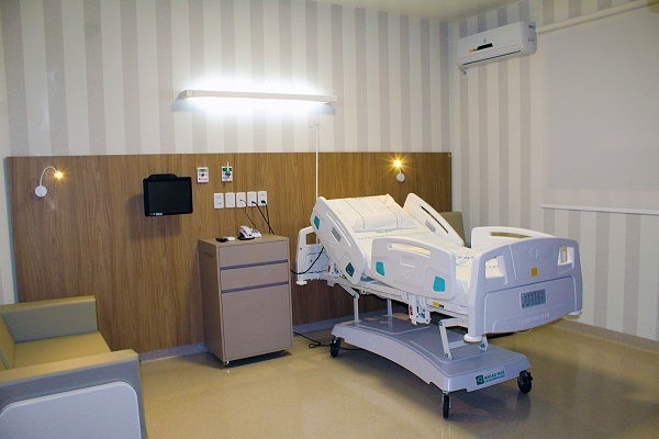 Imagem do quarto executivo. Na foto, aparece uma cama hospitalar com um móvel e um monitor ao lado. No lado esquerdo da foto, aparece um sofá.