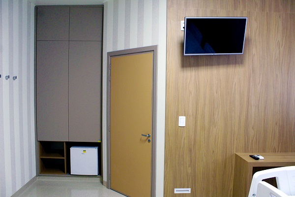 Imagem: detalhe do quarto executivo. No lado esquerdo, aparece um guarda-roupa embutido. Ao centro, temos a porta do banheiro fechada. No lado direito, aparece foto de uma parede com um televisor acima.