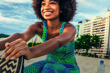 Foto de uma mulher sorrindo, fazendo alongamento na areia da praia com uma garrafa de água ao lado