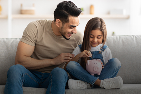 Imagem de um pai e uma filha colocando moeda em um cofre de porquinho
