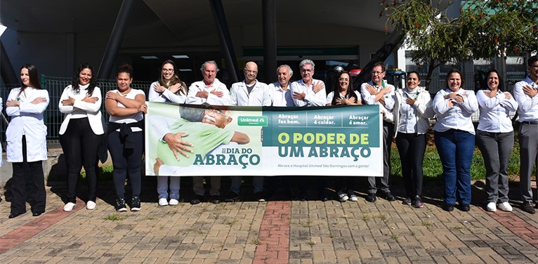 Na imagem, um grupo de pessoas está com os braços cruzados, simulando um abraço em si mesmo. As pessoas do centro seguram um banner com a frase "O poder de um abraço".