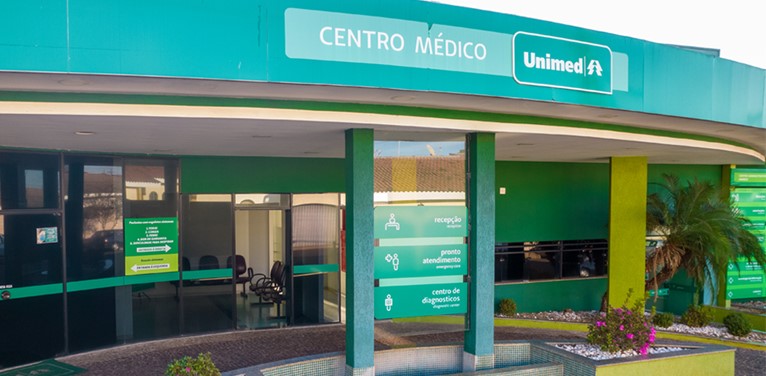 Imagem de um centro médico verde com uma árvore ao lado direito e portas de vidro.