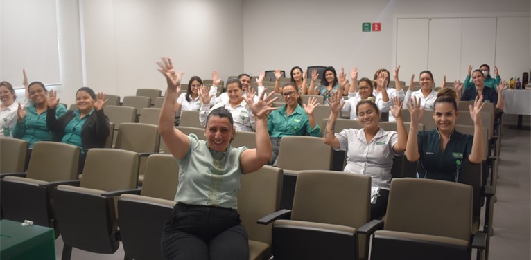 Na imagem, várias mulheres estão sentadas em um auditório, com as mãos para cima e sorrindo.