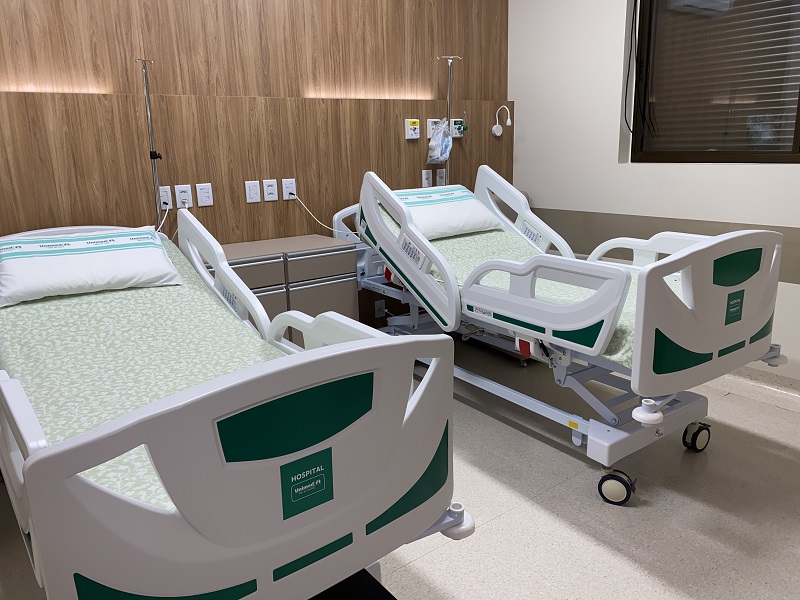 Imagem: Cama arrumada em um dos quartos de internação do Hospital Unimed Vale do Caí. Em cima dessa cama, está um travesseiro branco com o logotipo da Unimed e uma toalha branca com um kit de higienização.